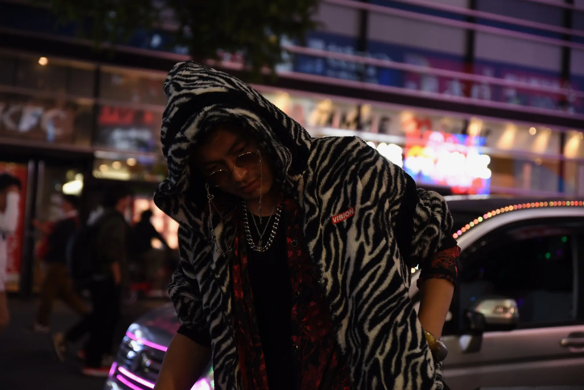 A person wearing sunglasses and a zebra-striped hooded coat stands beneath neon lights on a Shinjuku street at night.